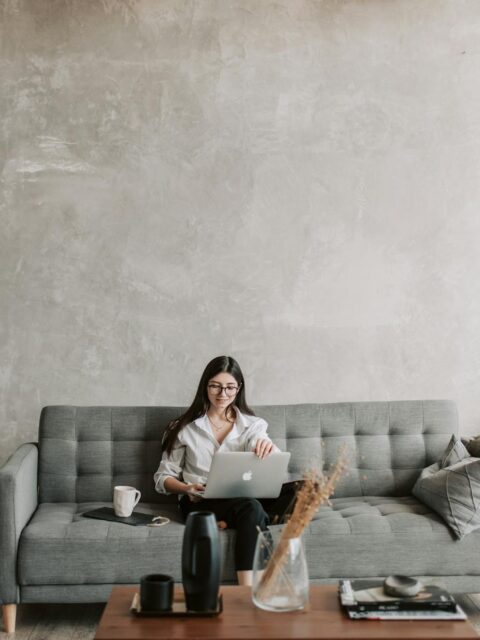 Happy female remote worker in eyeglasses sitting on cozy sofa with netbook and cup of coffee in loft style room with wooden floor and concrete wall